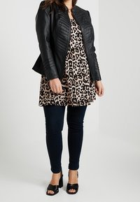 Zwarte faux leren jas met geribbeld detail, gedragen over een jurk met luipaardprint, gecombineerd met donkere skinny jeans en zwarte platformhakken.