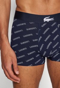 Bóxer en azul marino con un patrón repetido del logo en blanco "LACOSTE", con una cinturilla suave y una textura lisa.