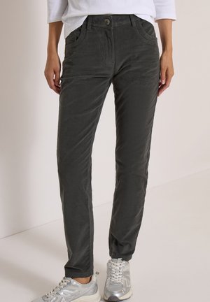Personne portant un pantalon en velours côtelé gris foncé et des chaussures de sport argentées, debout devant un fond uni.