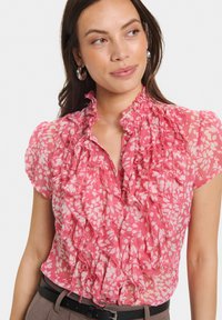 Pink floral blouse with ruffles along the front and neckline, short sleeves, and a lightweight, sheer fabric. Accented with buttons.