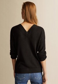 Schwarzer Pullover mit V-Ausschnitt, weichem Material, umgeschlagenen Ärmeln und geripptem Saum, kombiniert mit blauen Jeans.