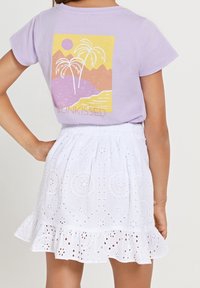 Fialové tričko s krátkým rukávem s grafikou palem, hor a slunce. V páru s bílou sukní s vyšívanými vzory a volánkovým lemem.