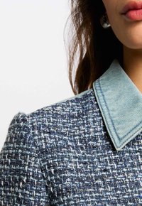 Niebieska marynarka z tweedu z jasnoniebieskim kołnierzem z denimu. Teksturowana tkanina ma wzór tkany z akcentami metalicznej nici.