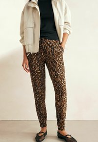 Leopard-Print-Jogginghosen aus braunem und schwarzem Stoff, mit einem elastischen Bund und Seitentaschen. Kombiniert mit einer hellbeigen Jacke.