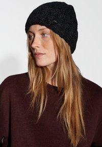 Black knitted beanie with a textured pattern; features a subtle logo tag. Worn with a dark maroon sweater, highlighting the hat's design.