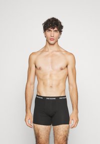Schwarze Baumwoll-Boxershorts mit einem Logo-Gummibund, die eine glatte Textur und ein tailliertes Design aufweisen, präsentiert von einem shirtlosen Modell vor einem hellen Hintergrund.