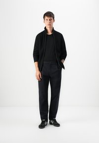 Felpa nera con zip e texture a coste, indossata sopra a una maglietta nera. Abbinata a pantaloni scuri e sneakers nere con strisce bianche.