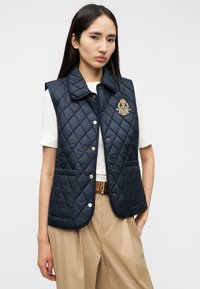 Μπλε ναυτικό γιλέκο με καπιτονέ ύφασμα, γιακά, κουμπιά τύπου σναπ και μπροστινές τσέπες, με κεντημένο έμβλημα στο αριστερό μέρος του στήθους.