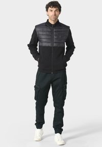 Chaqueta negra con pecho acolchado, cierre delantero con cremallera, cuello alto; combinada con pantalones cargo oscuros y zapatillas blancas. Diseño y textura casual y deportiva.