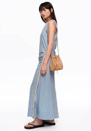 Mujer con vestido largo sin mangas azul a rayas y chanclas negras, lleva un bolso tipo cubo marrón con estampado de palmeras al hombro.