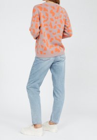 Strickpullover in Hellpink mit kräftigen orangefarbenen abstrakten Mustern, kombiniert mit hellblauen Jeans und weißen Turnschuhen.