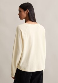 Long-sleeve crewneck shirt in soft, light cream fabric with textured pattern; features raglan sleeves and a relaxed fit with a curved hem.