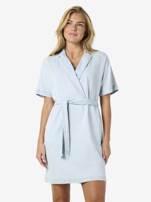 Mujer con vestido cruzado con cinturón de manga corta en azul claro, de pie contra un fondo blanco liso, mirando ligeramente hacia arriba.