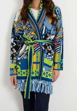 Langer bunter Cardigan mit abstrakten Mustern und Sternen, tailliert mit Gürtel und Fransen am Saum, getragen über dunkler Hose und weißem Oberteil.