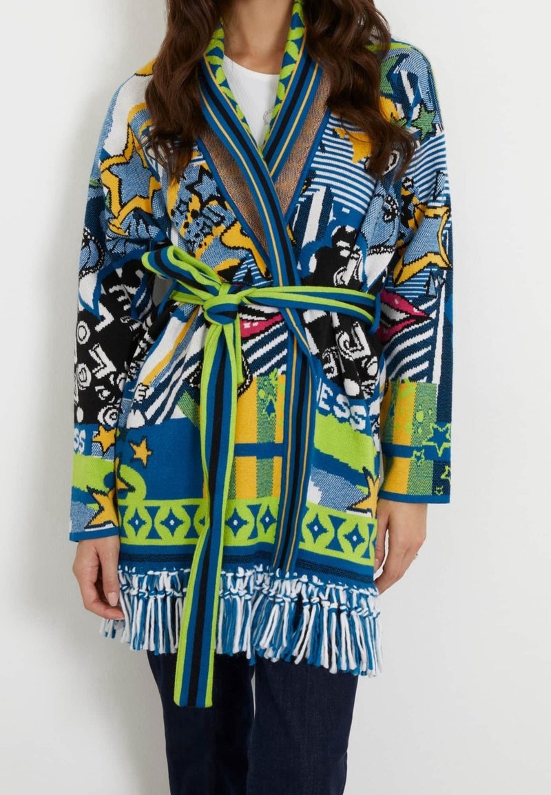 Long gilet coloré avec des motifs abstraits et des étoiles, taille ceinturée et ourlet à franges, porté sur un pantalon foncé et un haut blanc.