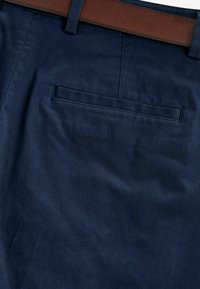 Vue de dos d’un pantalon bleu marine avec une poche passepoilée unique et une ceinture en cuir marron passée dans les passants.
