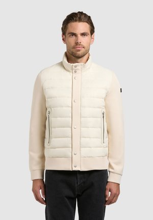 Mees kannab kreemjat värvi polsterdatud jopet, millel on trukid ja lukuga taskud, seisab ühevärvilise heleda tausta ees.