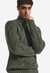 Mörkgrön kabelstickad polotröja med ribbade ärmslut och nedkant. Har ett strukturerat mönster med tjockt, varmt material.