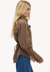 Chemise marron à carreaux avec un détail à nouer dans le dos, manches longues et col, associée à un jean taille haute en denim.