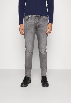 Mann trägt schlank geschnittene, ausgebleichte graue Jeans, schwarze Schnürstiefel und ein dunkelblaues Langarmhemd, steht vor einem schlichten hellen Hintergrund.