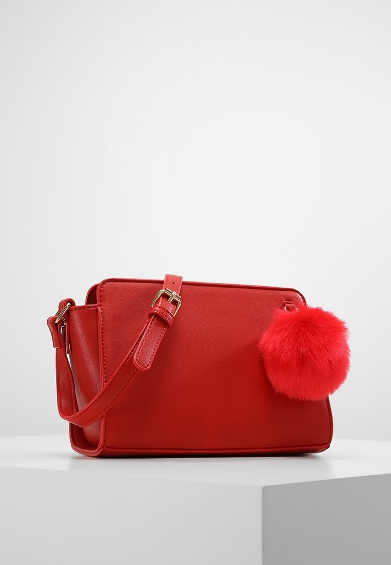 Czerwona torebka na ramię z eko skóry z regulowanym paskiem, prostokątnym kształtem, gładką fakturą oraz puszystym czerwonym pomponem jako ozdobą.