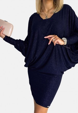 Kvinde iført en glitrende navyblå kjole med flagermusærmer, der holder en beige clutch og er accessoriseret med et ur, en ring og en halskæde.