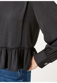 Maglietta nera realizzata in tessuto testurizzato, con vita peplum, maniche lunghe e polsini con bottone. I dettagli includono una finitura morbida e arricciata.