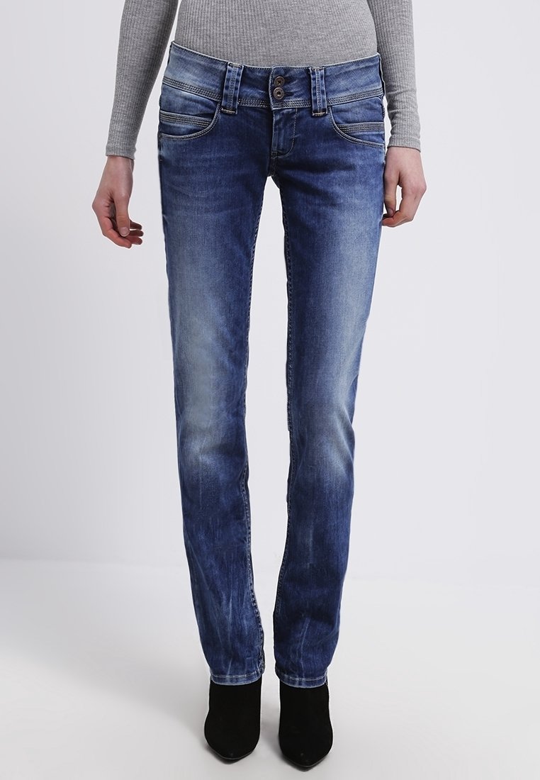 Person som har på sig urtvättade blå skinny jeans, en grå ribbad långärmad topp och svarta spetsiga stövlar, som står mot en enkel bakgrund.