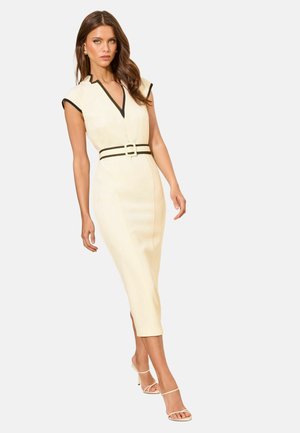 Žena v priliehavých krémových šatách bez rukávov s čiernym lemom a opaskom, v kombinácii s krémovými vysokými sandálmi, stojaca na jednoliatom pozadí.