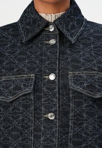 Jakke i denim med mørkeblå, mønstrede tekstiler. Har krage, sølvfargede knappene og lommer som fremhever intrikate design.