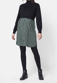 Czarny sweter z kołnierzem typu mock turtleneck, zestawiony z zieloną sukienką w czarne kropki, z guzikami z przodu i luźnym krojem w talii.