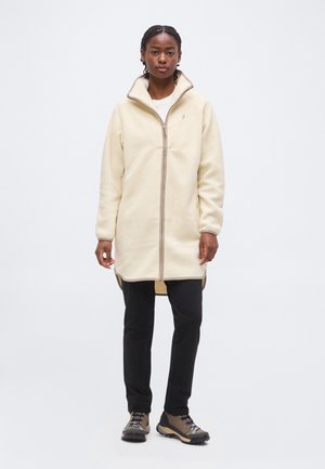 Femme portant une longue veste polaire crème zippée, un pantalon noir et des chaussures de randonnée marron, debout devant un fond clair uni.