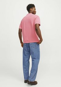 Roza polo majica s kratkimi rokavi, sproščenega kroja, v kombinaciji s svetlo modrimi kavbojkami in rjavimi čevlji, gledano s hrbta.