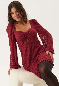 Robe en dentelle bordeaux avec un décolleté carré, des manches bouffantes et un corsage froncé. Possède un ourlet à volants et un motif texturé.