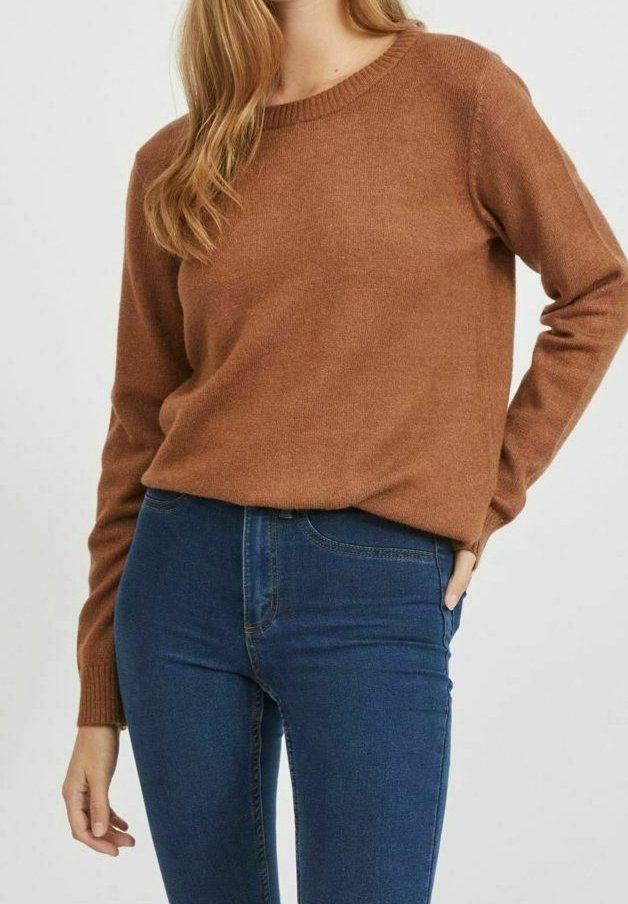 Femme portant un pull à manches longues marron et un jean taille haute bleu, debout avec une main dans la poche devant un fond uni.