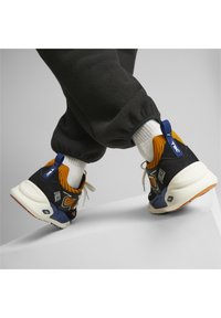 Baskets avec une tige en suède noir et en maille, accents orange, semelle intermédiaire blanche et semelle en caoutchouc. Présente un logo bleu sur le talon.