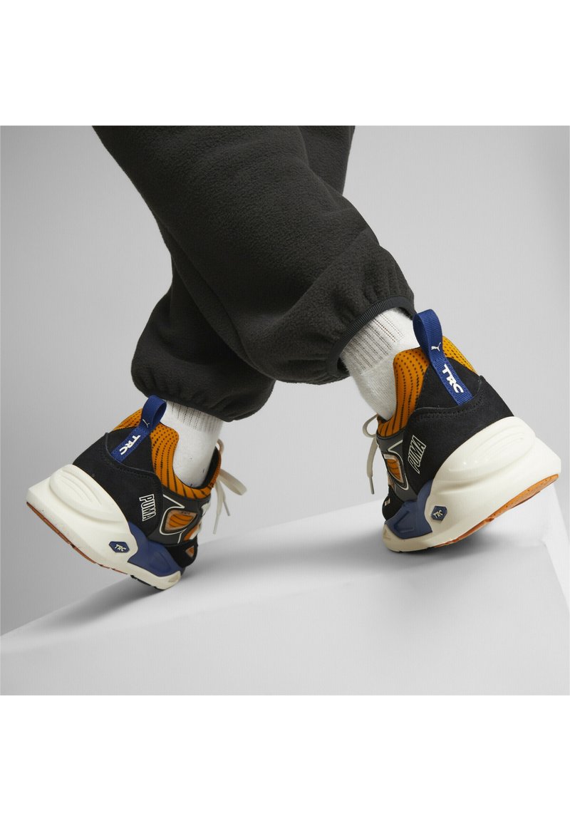 Baskets avec une tige en suède noir et en maille, accents orange, semelle intermédiaire blanche et semelle en caoutchouc. Présente un logo bleu sur le talon.