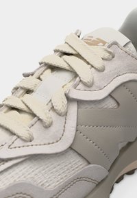 Gros plan sur une chaussure de sport beige avec des lacets texturés, des superpositions en suède, des panneaux cousus et un tissu côtelé sur la partie avant supérieure.