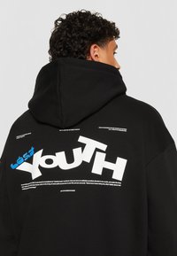 Sort hættetrøje med stort hvidt "Youth" tekst og blå accenter; har sparsomme grafiske tekstdetaljer på ryggen; materiale af bomuldsblanding.