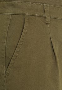 Primo piano di pantaloni in tessuto verde oliva che mostra la tasca anteriore, il passante della cintura e la cucitura dettagliata.