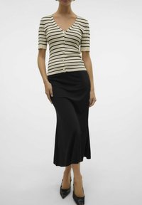 Prugasta bež in črna kratka kardigan s kratkimi rokavi in gumbi, v kombinaciji s prilegajočo črno maksi krilo, stilizirano s koničastimi črnimi čevlji.