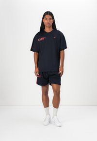 Navy mesh t-shirt with red logo accents, paired with navy shorts. Outfit features a relaxed fit and lightweight fabric. White sneakers and socks.