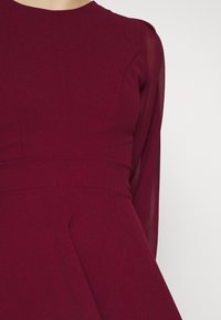 Gros plan d'une robe rouge profond avec corsage ajusté et manches longues transparentes, montrant la texture du tissu et les détails des coutures sur un fond clair.