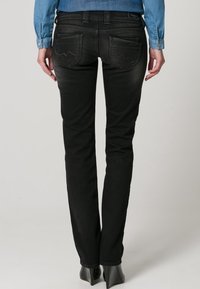 Jeans de mezclilla negra con corte ajustado, que cuentan con bolsillos traseros con costuras en forma de ola y una cintura de altura media. La textura se ve suave y uniforme.