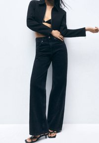 Giacca nera corta con zip, abbinata a jeans neri a vita alta e gamba larga e sandali neri con cinturini. Tessuto liscio e linee pulite.