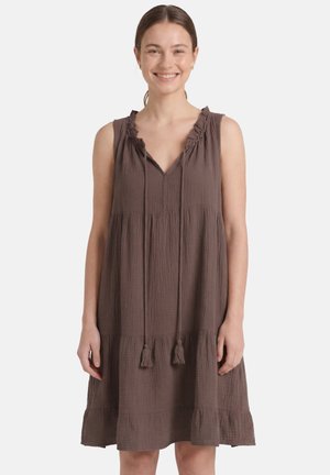 Femme souriante aux cheveux bruns attachés, portant une robe marron à volants sans manches avec des liens à pompons au niveau d'un col à volants.