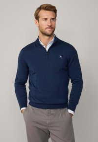 Suéter de punto azul marino con cuello de media cremallera, puños acanalados y dobladillo. Presenta un pequeño logotipo bordado en el lado izquierdo del pecho. Llevado sobre una camisa con cuello.