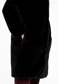 Sort pelsjakke med blød tekstur, lange ærmer og sidelommer, i en afslappet pasform der når til midt på låret.
