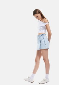 Hellblauer Denim-Wickelrock mit Bindegürtel in der Taille, kombiniert mit einem weißen Off-Shoulder-Crop-Top und weißen Sneakers mit Schnürdetails.