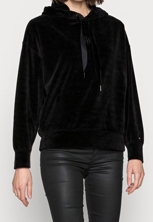 Femme portant un sweat à capuche en velours noir avec cordons et un pantalon en cuir noir ajusté sur un fond uni.